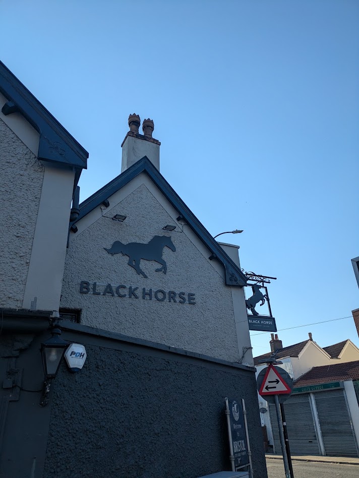 A shaded wall with the Black horse sign and logo on, with the hanging sign visible too against a blue sky