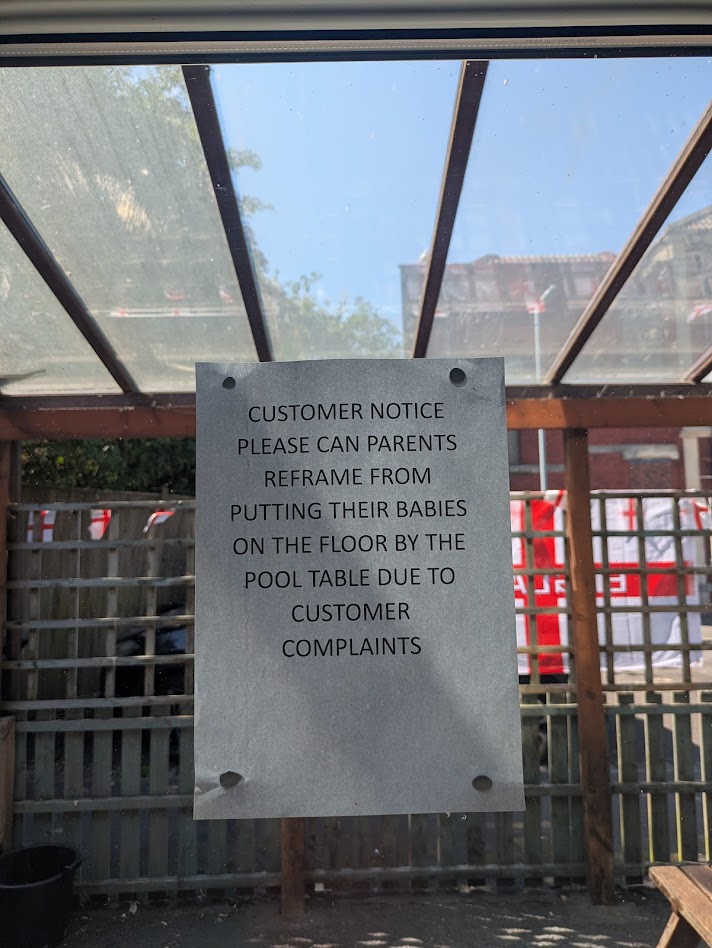 A printed sign stuck on the window reading in all caps 'CUSTOMER NOTICE PLEASE CAN PARENTS REFRAME [sic] FROM PUTTING THEIR BABIES ON THE FLOOR BY THE POOL TABLE DUE TO CUSTOMER COMPLAINTS