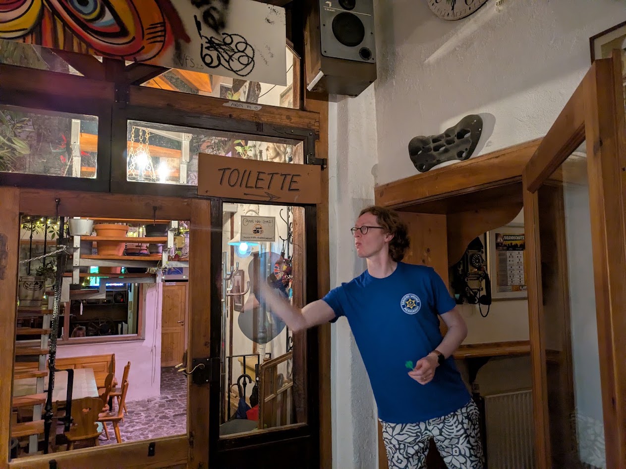 A shot of Jrat throwing a dart. There's a sign above his head saying toilette