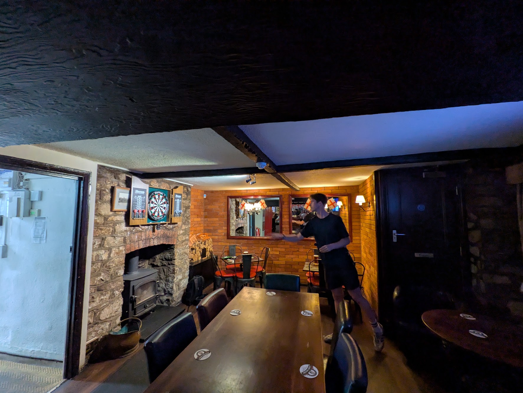 A wide shot of the oche, showing a low ceiling with Sam throwing darts, and quite a dense collection of tables
