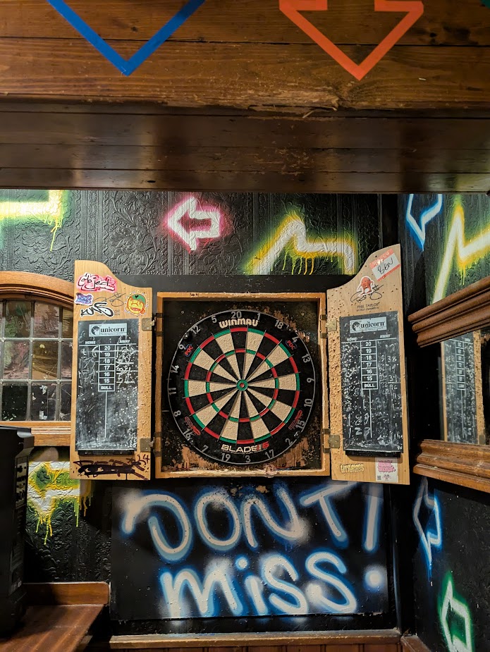 A picture of the dartboard in the Bell. The board is in decent nick, but the cupboard it's in is riddled with quite large holes from very wide of the mark arrows. There is a beam above the oche which also has significant dart damage