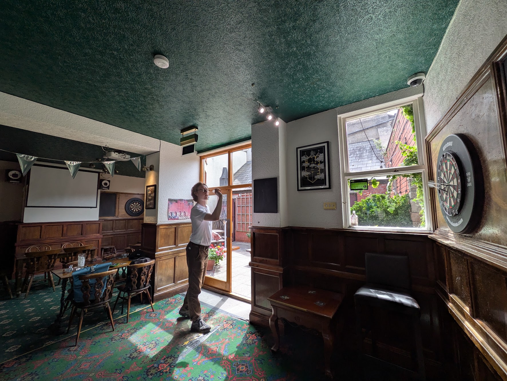 A wider shot of a ratty looking man throwing darts on the Oche