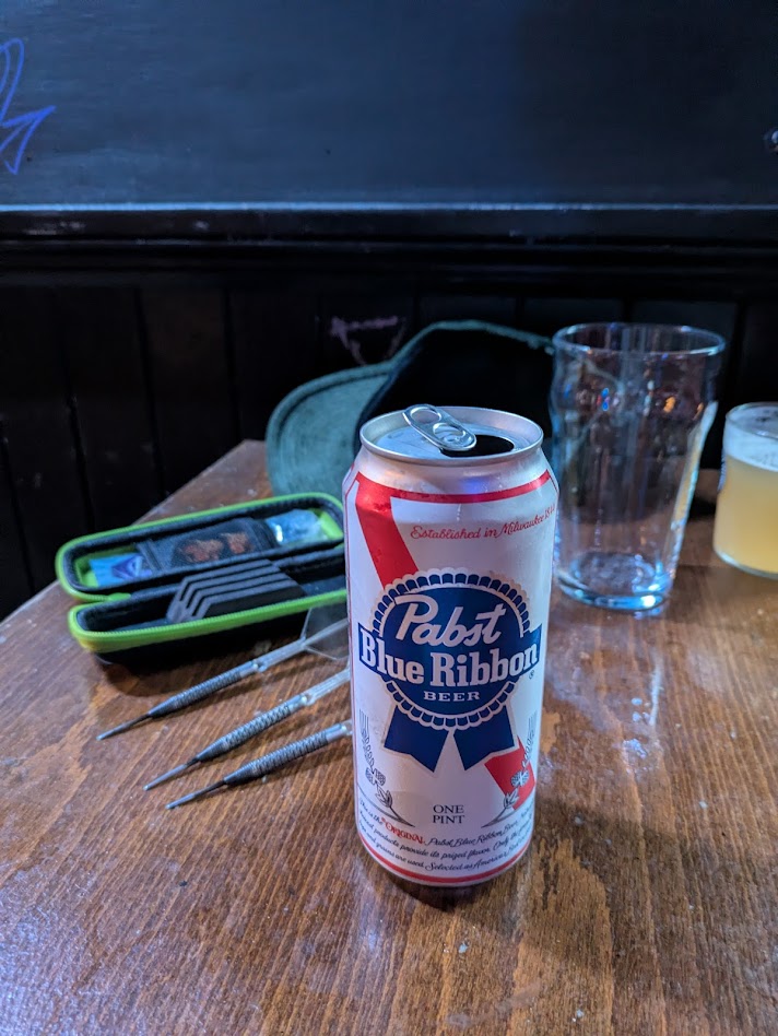 A can of Pabst Blue Ribbon sat on a table, my darts are in the background